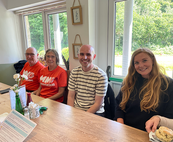 two mnd volunteers wearing orange mnd t-shirt sitting at a table with Alys Griffiths and Dr Louis Stokes